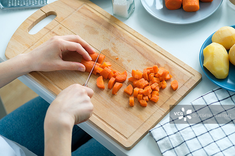 一个女人把煮熟的胡萝卜切成方块。准备一种蔬菜沙拉，叫做油醋酱。菜刀放在菜板上。附近还有其他食材。素食和纯素食品的概念。图片素材