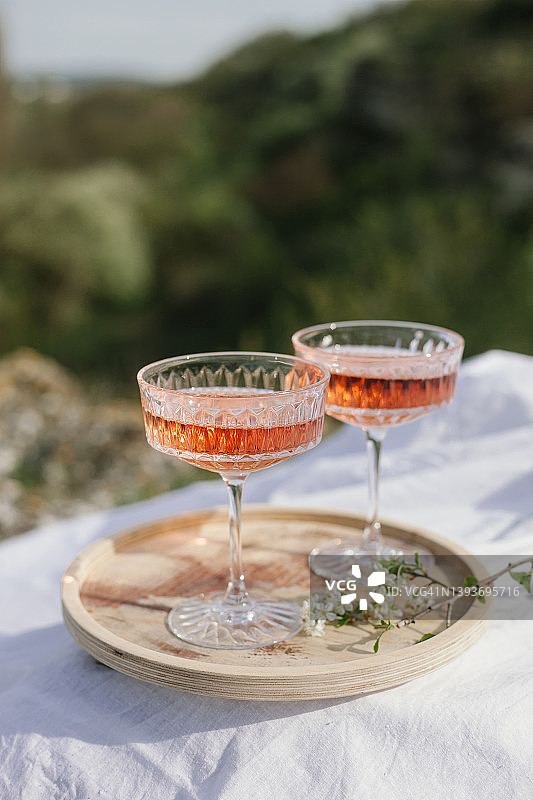 两杯玫瑰红葡萄酒放在木制托盘和白色纺织品上，在春天野餐。阳光明媚的日子，绿草如茵，樱花盛开。图片素材