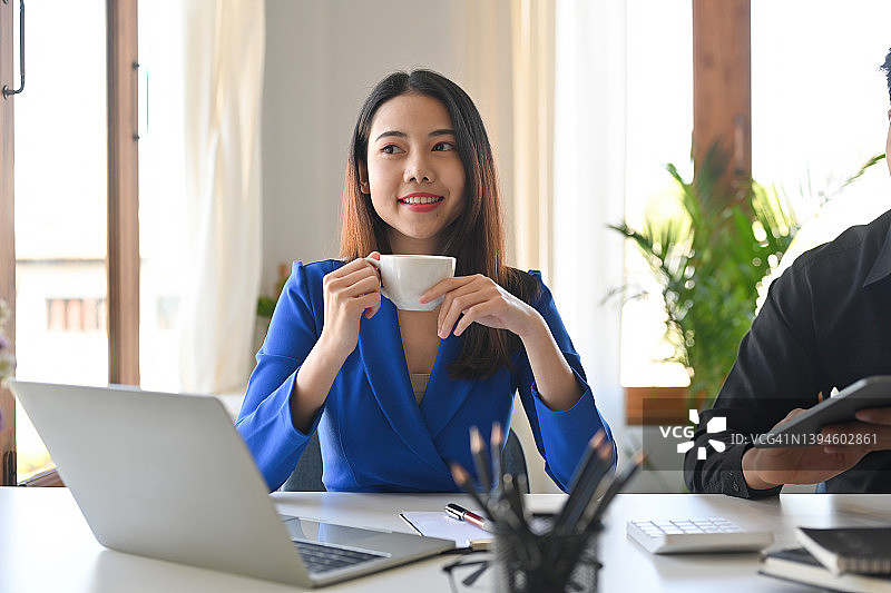 一个年轻的微笑漂亮的亚洲商业女性的正面视图肖像看向一边，拿着一杯咖啡在办公室的笔记本电脑上工作，为商业和技术概念。图片素材