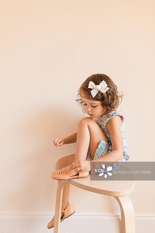 3岁蹒跚学步的小女孩穿着柔和的连衣裙和白色蝴蝶结，坐在椅子上穿鞋图片素材