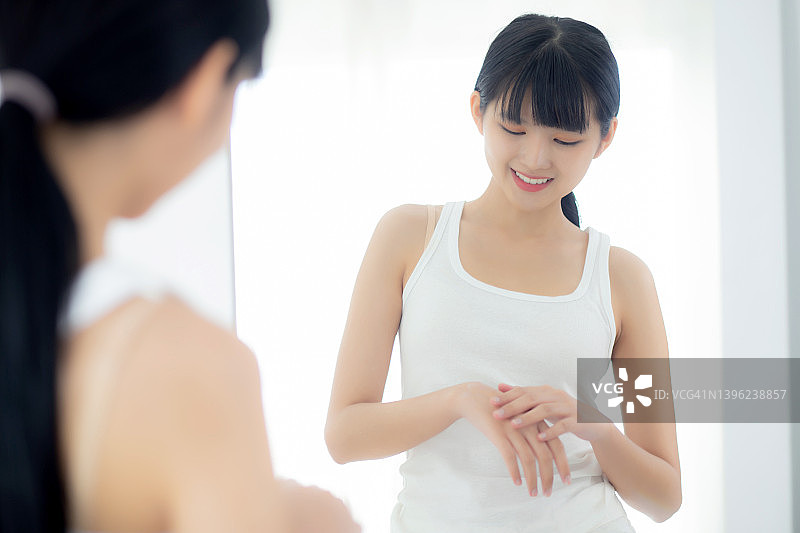 美丽的年轻亚洲女性看着镜子，并应用润肤霜和乳液与手的皮肤保护，皮肤护理和美容美容，治疗和身体护理，皮肤护理的概念。图片素材