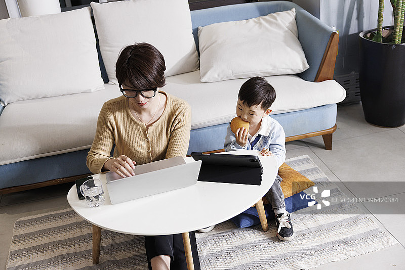 年轻女性在家里用笔记本电脑工作，她们的儿子在旁边玩图片素材