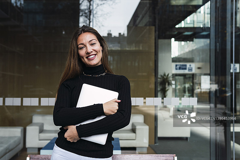 微笑的年轻女商人拿着笔记本电脑站在玻璃墙前图片素材