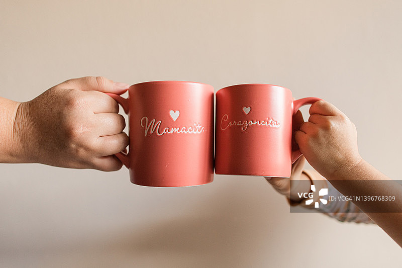 一个母亲和女儿的手拿着粉红色的杯子，上面写着“Mamacita”和“Corazoncita”，西班牙语是可爱的“妈妈”和“小爱”的意思。图片素材