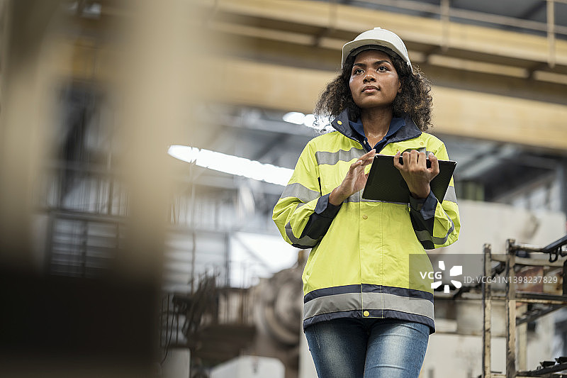 打造制造业质量安全文化。低角度视角，一名女性非洲裔美国工程师手持数字平板电脑，站在工厂周围的机器。图片素材