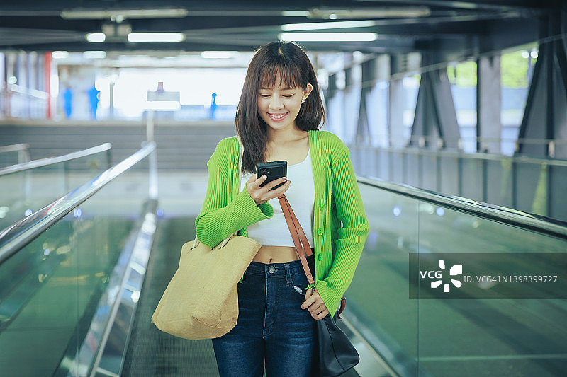 年轻的亚洲女商人在机场的自动扶梯上用手机阅读电子邮件。商务旅行的概念。图片素材