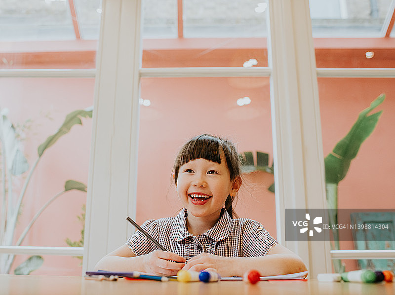 一个可爱、快乐的小女孩坐在桌子旁，带着灿烂的笑容，用一支铅笔完成作业图片素材