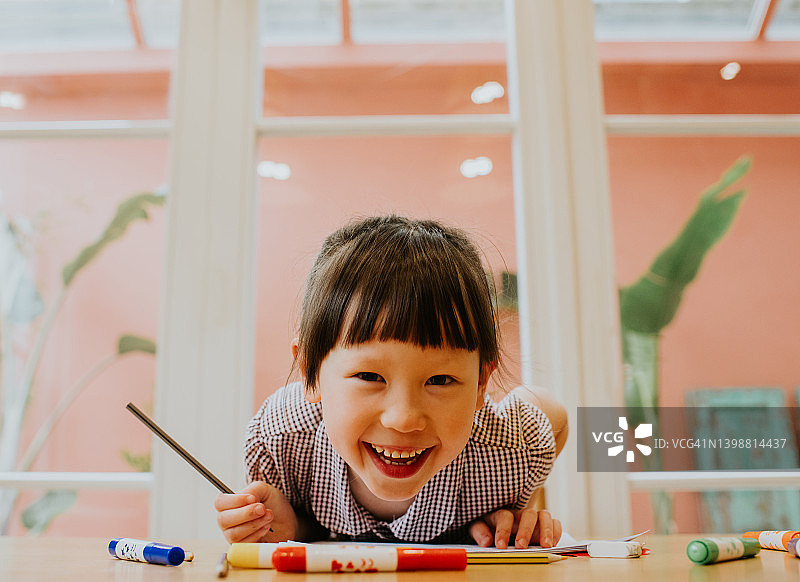 一个可爱、快乐的小女生斜靠在桌子上，面带灿烂的笑容，直视着镜头。她握着一支铅笔。图片素材