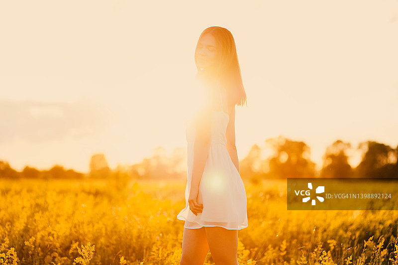 快乐的年轻女子，深色长发，穿着白色的衣服，微笑着站在草地上，在明媚的阳光明媚的日子里图片素材