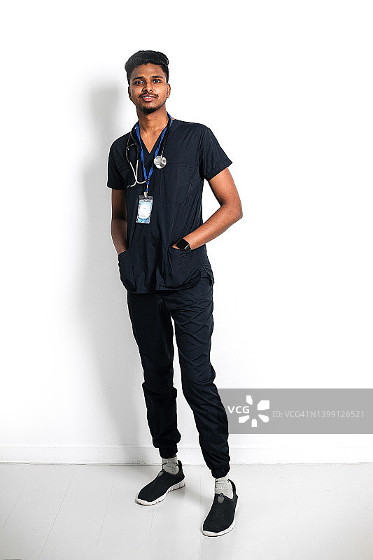 微笑的印度医生穿着医用棉衣，拿着听诊器站在诊所的白墙上。图片素材