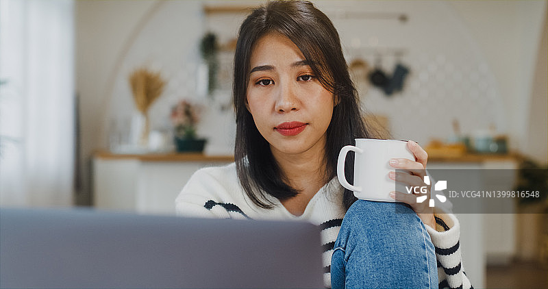 自由亚洲女士穿着休闲，使用笔记本电脑，在家里的厨房喝咖啡。图片素材