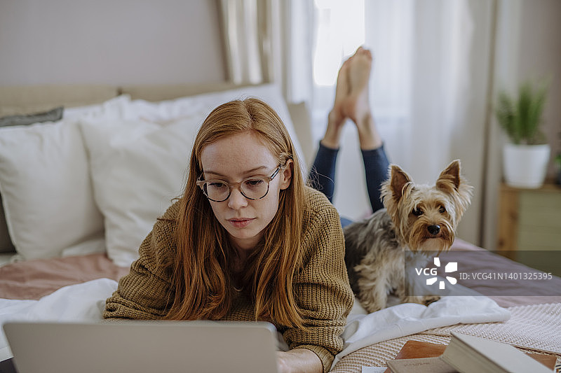 戴眼镜使用笔记本电脑的女人躺在家里的床上的宠物狗图片素材