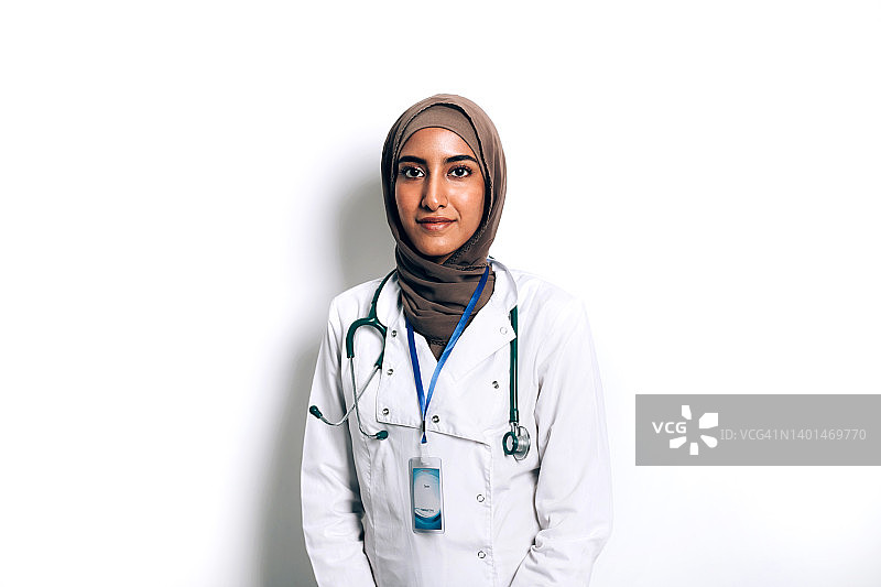 微笑的亚裔穆斯林女医生身穿白大褂，戴着头巾，拿着听诊器，靠在诊所的白墙上。图片素材