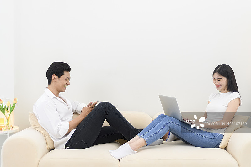 微笑的年轻夫妇在家里的客厅使用笔记本电脑和手机图片素材