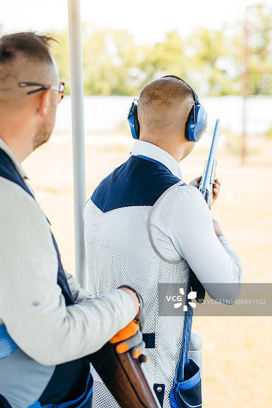 两个戴着墨镜，戴着防护耳机，穿着步枪背心的成年男子正在练习射击。图片素材
