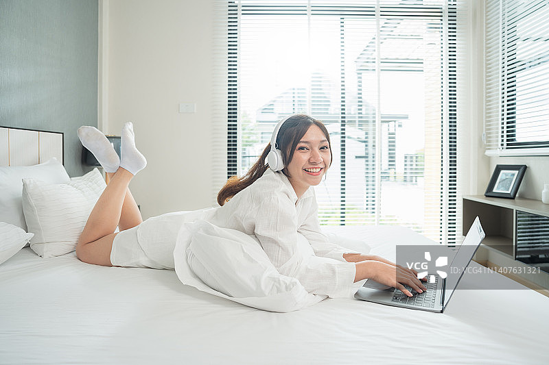 美丽的年轻女子戴着耳机在卧室里放松。年轻女性躺在床上使用笔记本电脑。自由职业者。在家里工作。在线教育图片素材