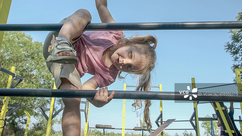 小女孩在户外运动场上爬体操梯。阳光灿烂的日子里，可爱的小女孩爬上城市公园里的垂直运动梯。图片素材