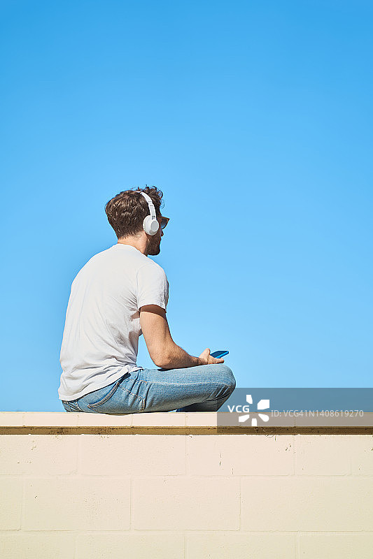 年轻人穿着休闲的衣服，坐在以蓝天为背景的黄色砖墙上，一边用手机一边戴着无线耳机听音乐。图片素材