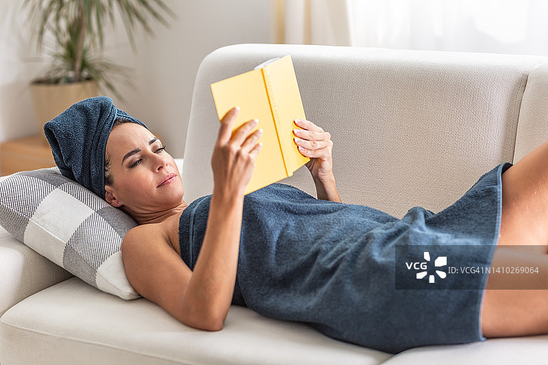一位裹着蓝色毛巾的美女正在家里的沙发上看书。图片素材