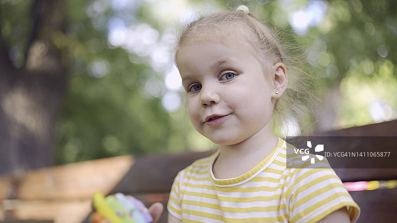 小女孩拿着一个五颜六色的姜饼微笑着。特写肖像可爱的孩子女孩坐在公园的长椅上饼干在看着相机镜头。图片素材