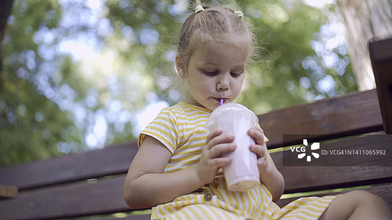 小女孩用吸管喝奶昔。特写可爱的小女孩坐在公园的长凳上喝奶昔图片素材