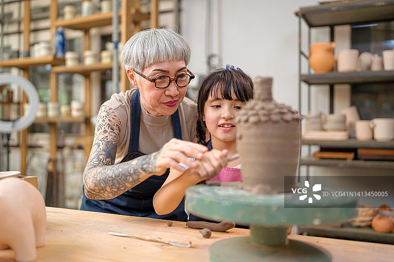 高级女艺术家教年轻女孩在陶工的轮子上创造陶器。图片素材