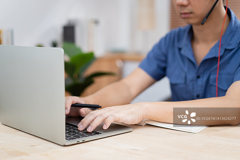中年亚洲男性，戴着耳机，坐在家庭办公室，用笔记本电脑为客户提供呼叫中心支持图片素材