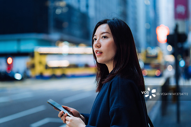 在繁忙的城市交通和现代摩天大楼的背景下，专业年轻的亚洲女商人在市中心的城市街道上使用智能手机上下班。商业与技术的概念图片素材