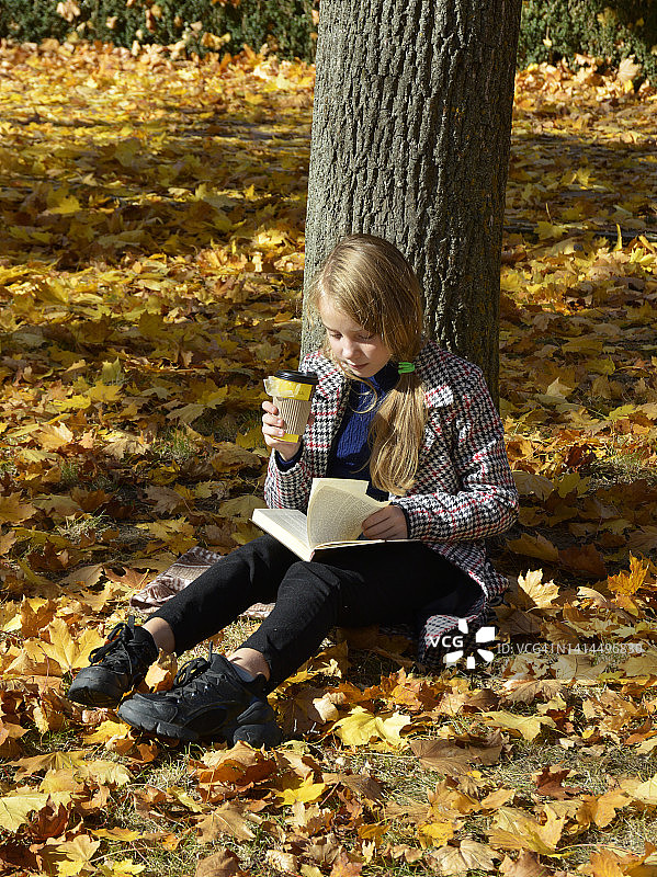 秋天的公园里，一个女孩拿着一本书和一杯咖啡坐在落叶上图片素材