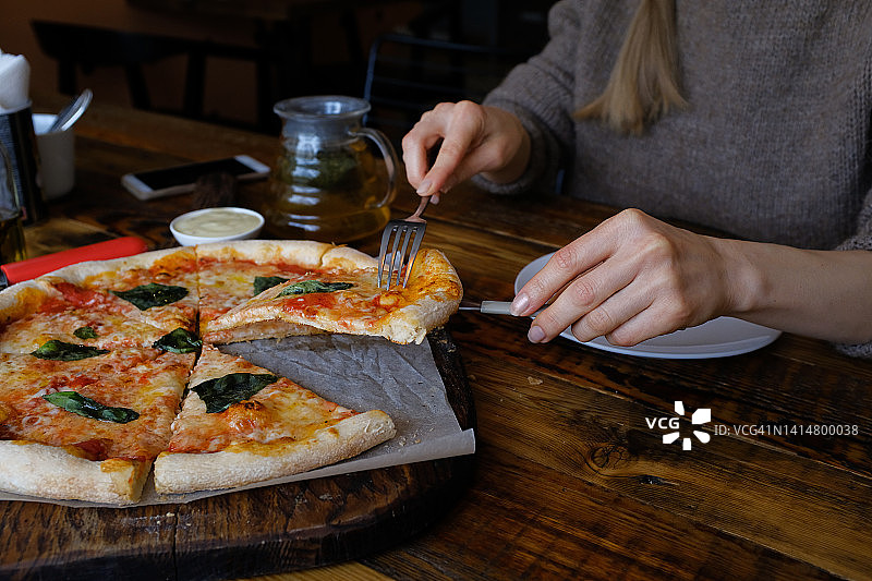 一位女士在咖啡馆或快餐店的披萨馆里用马苏里拉奶酪、西红柿、罗勒切一块素食披萨。美味的意大利美食。切成薄片的玛格丽塔披萨放在木板上或摆盘架上。不健康的午餐、点心。图片素材