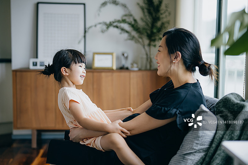 可爱的亚洲小女孩坐在妈妈的腿上，在家里放松的时候和妈妈聊天。享受母女的时刻。家庭生活方式。爱与关怀的理念图片素材