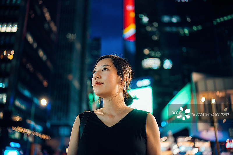 自信的亚洲女商人以乐观的表情仰望，站在繁华和照明的市中心街道与繁忙的交通和高耸的企业摩天大楼在晚上。女性领导力。商业成就和成功图片素材
