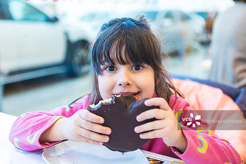 穿着粉色毛衣的小女孩在自助餐厅，吃着巧克力松饼，嘴巴和手都很脏。图片素材