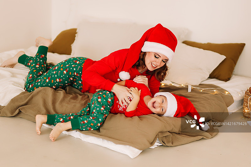 快乐的妈妈和女儿穿着红色睡衣，戴着圣诞老人的帽子，在舒适的卧室里开心地拥抱和装饰圣诞树。等待新年的到来。圣诞拍摄的概念。一个有一个孩子的家庭正在为平安夜做准备图片素材