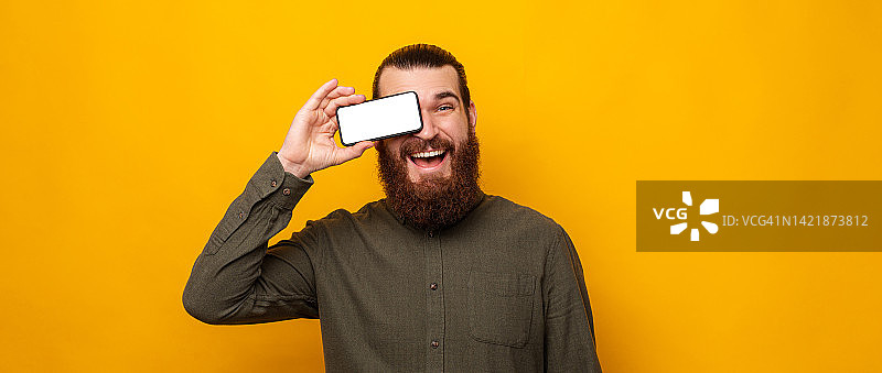 顽皮的大胡子男子用手机屏幕遮住一只眼睛，对着镜头微笑。图片素材
