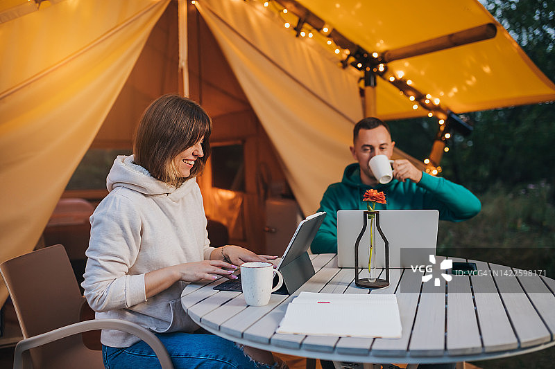 幸福的家庭夫妇自由职业者在一个舒适的野营帐篷在夏天的晚上使用笔记本电脑。户外度假度假的豪华露营帐篷。生活方式的概念图片素材