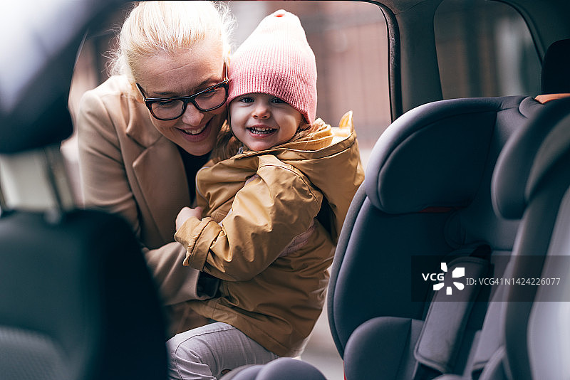 快乐的母亲把她的小女儿放在汽车安全座椅上的画像图片素材