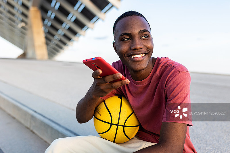 快乐的年轻黑人男子带着篮球在户外放松，同时用智能手机发送音频语音信息-科技，青年和千禧一代的人的概念图片素材