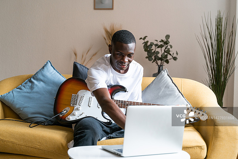 一名非洲裔美国男子在家中客厅使用笔记本电脑，在他的笔记本电脑上寻找弹奏吉他的乐谱。图片素材