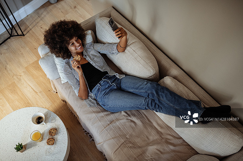 微笑的非裔美国妇女放松在沙发和看智能手机在客厅图片素材