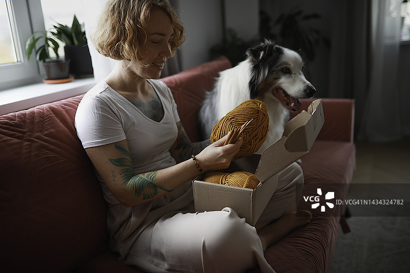 可爱的女人在舒适的客厅和狗狗在沙发上打开纱线包裹的盒子。图片素材