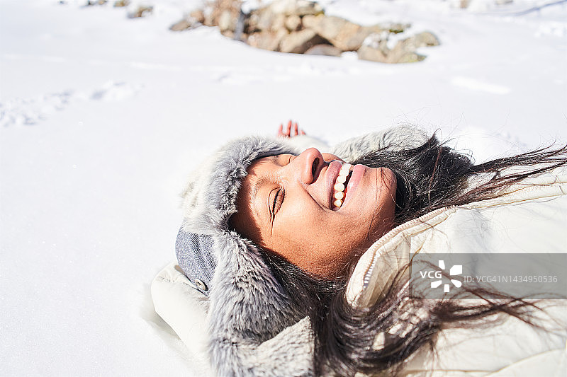 一位拉丁美洲的年轻女士躺在雪地上，闭着眼睛微笑。图片素材