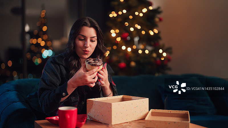 圣诞节期间，年轻女子打开礼物/订阅盒，享受吃甜甜圈，同时在家里的客厅放松图片素材