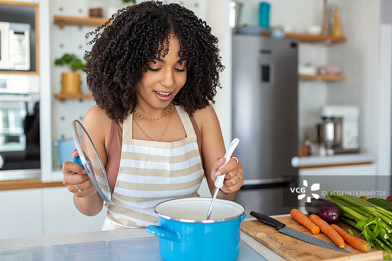 漂亮的非洲裔美国妇女拿着烹饪勺，用新鲜的蔬菜在厨房煮汤的照片图片素材