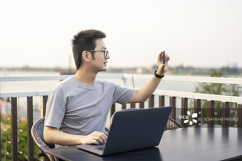 亚洲男性自由职业者在河边的露天咖啡馆用笔记本电脑和手机图片素材
