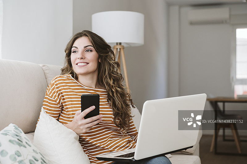 微笑的年轻女子与笔记本电脑和手机在家里的沙发上图片素材