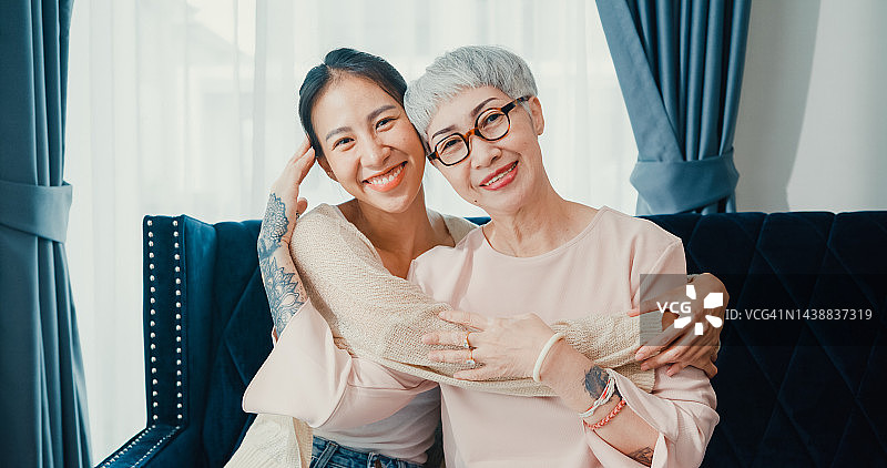 快乐年轻的亚洲女儿和退休的资深母亲坐在舒适的沙发沙发拥抱乐趣享受家庭时刻在家里。图片素材
