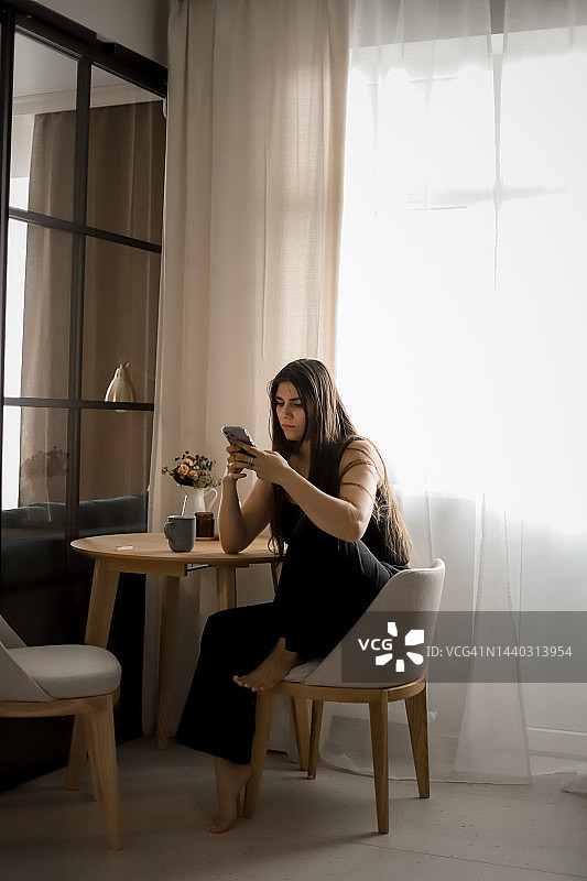 一个身穿深色衣服、头发飘逸的女孩坐在厨房的椅子上，用手机通话图片素材