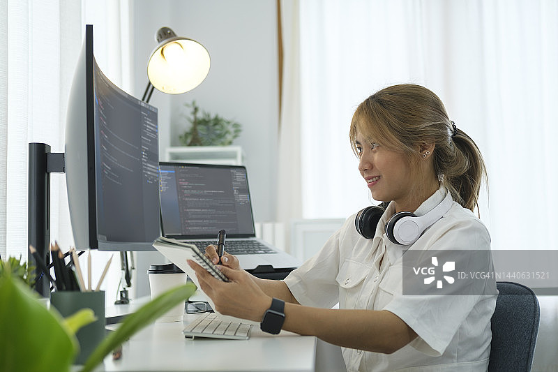 微笑的女程序员坐在巨大的曲面电脑显示器前，在现代的工作场所写程序代码。图片素材