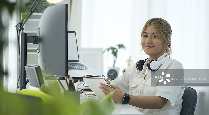 在白色现代办公室内部，微笑的女性IT开发人员坐在大曲面电脑显示器前的肖像。图片素材
